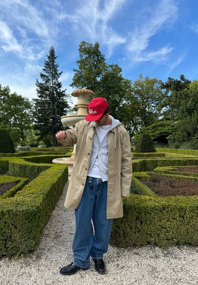 Impermeable beige sobre una sudadera gris, combinado con jeans azules sueltos y zapatos negros. Gorro de béisbol rojo y llavero visibles. Fondo de jardín.