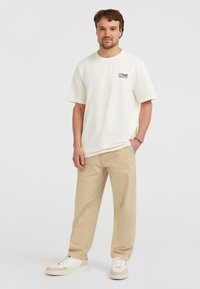 Licht beige oversized t-shirt met korte mouwen, voorzien van zwarte logo-print. Gecombineerd met licht kaki broek en witte sneakers.