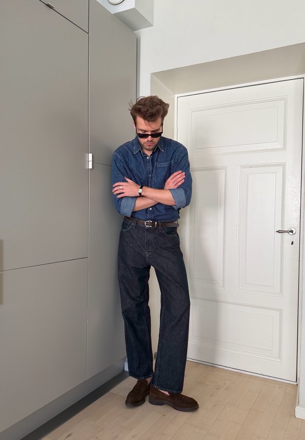 Mann mit Sonnenbrille, Jeanshemd und dunkler Jeans steht mit verschränkten Armen an eine Wand gelehnt nahe einer weißen Tür im Innenbereich.