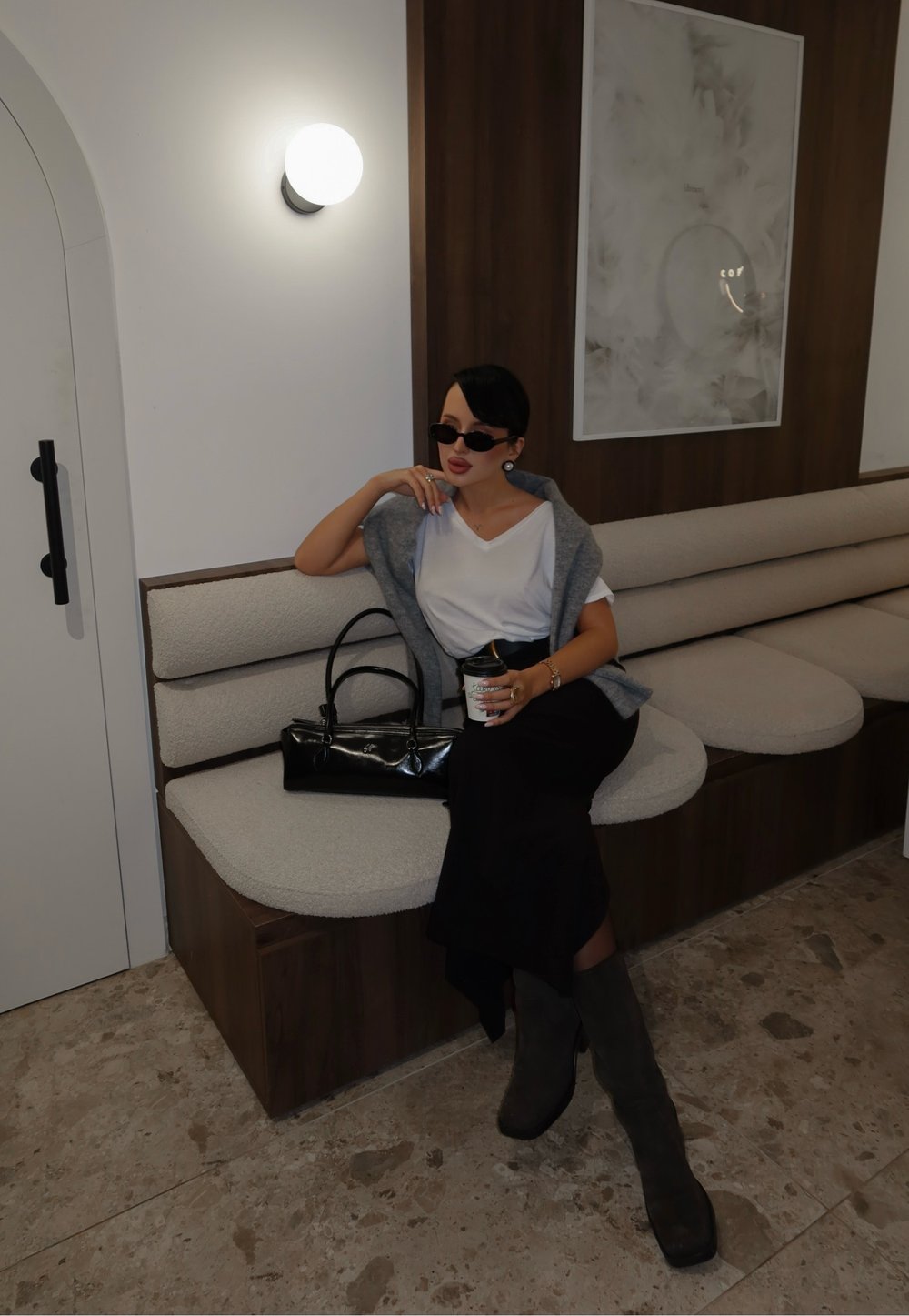 Femme en lunettes de soleil portant un haut blanc et une jupe noire, assise sur un banc rembourré, tenant une tasse de café dans un espace intérieur moderne.