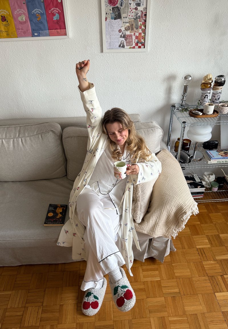 Peignoir blanc confortable avec petits motifs d'oiseaux, assorti à un pyjama blanc et des chaussons avec des accents cerise. Assis sur un canapé beige, tenant une tasse.