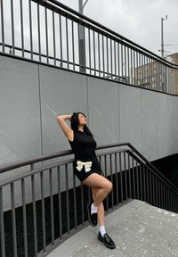 Haut noir et jupe, présentant un grand nœud blanc à la taille. Les accessoires comprennent des chaussures noires et des chaussettes blanches. Cadre urbain avec des murs gris.