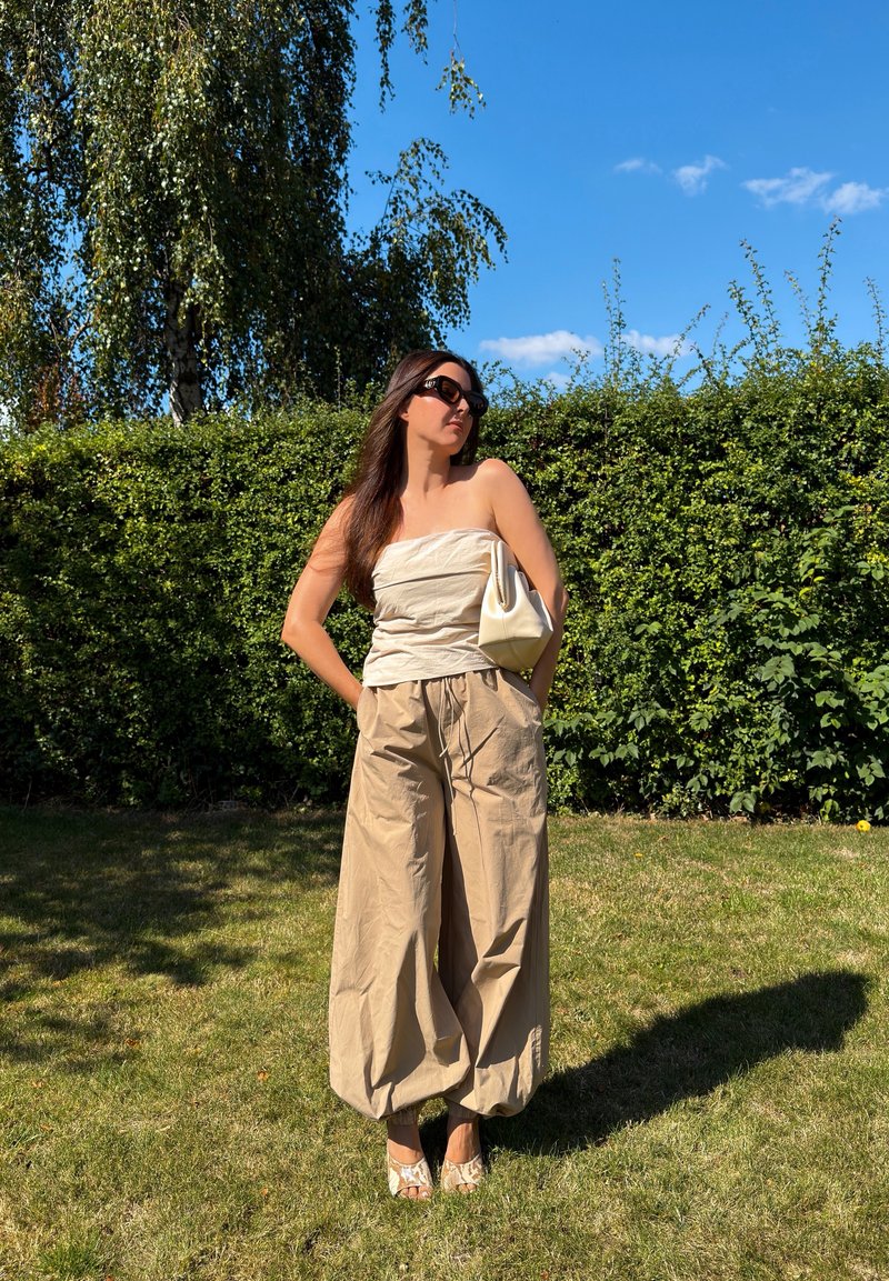Woman wearing beige strapless top and wide pants standing on grass with hedge and blue sky in the background, holding a white bag.