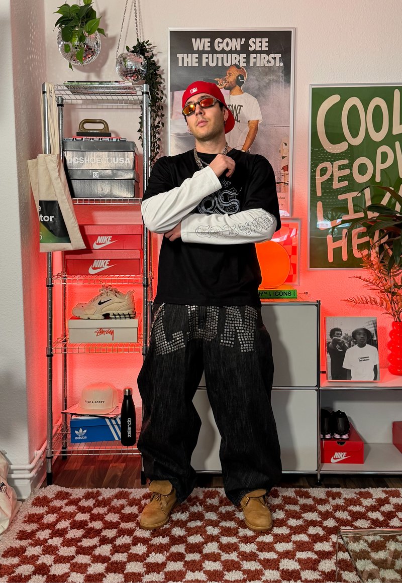 Homme portant une casquette rouge, des lunettes de soleil, une chemise à manches longues noire et blanche, et un pantalon noir clouté, se tenant dans une pièce avec des baskets, des affiches et des plantes.
