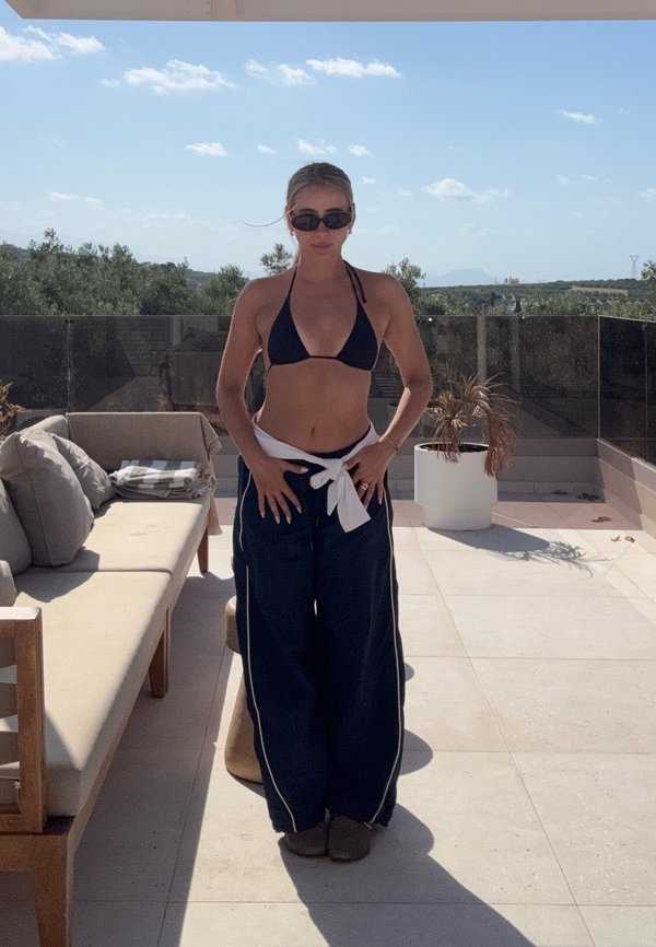 Sieviete melnā bikini augšdaļā un brīvās tumši zilās biksēs stāv saulainā terasē ar dīvānu un podā esošu augu zem skaidras debess.