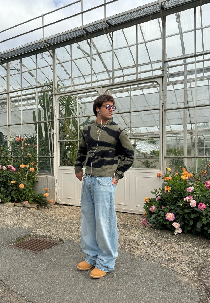 Πλεχτό πουλόβερ σε πράσινο και μαύρο καμουφλάζ, χαλαρές ανοιχτό γαλάζιες τζιν παντελόνες, μπεζ ξυλοπέδιλα, ασημένια κοσμήματα, με θερμοκήπιο και λουλούδια στο φόντο.