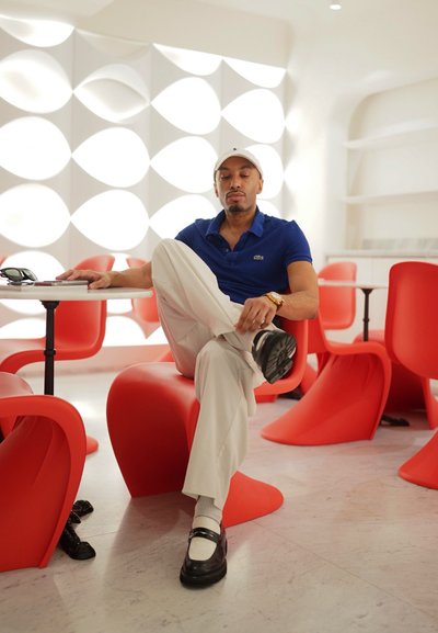Hombre sentado con una pierna cruzada en una silla roja en una habitación moderna con paneles de pared geométricos blancos y varias sillas rojas.