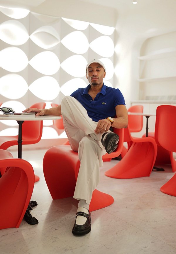 Hombre sentado con una pierna cruzada en una silla roja en una habitación moderna con paneles de pared geométricos blancos y varias sillas rojas.