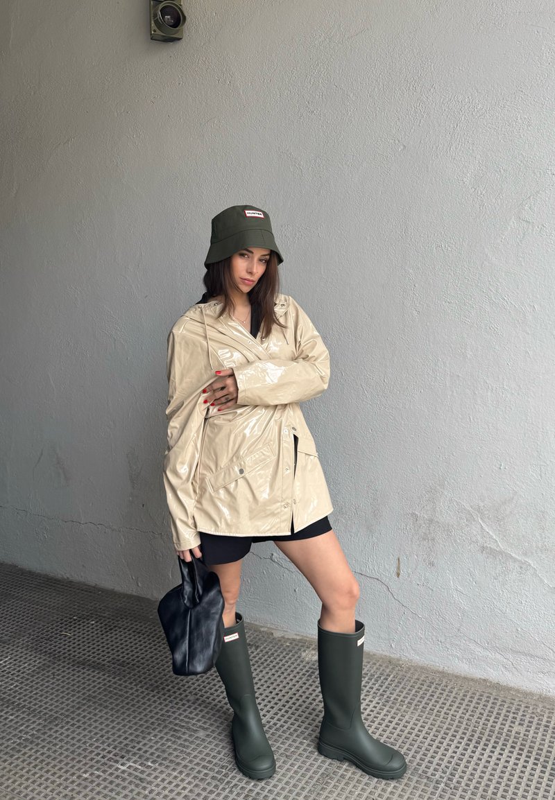 Femme portant un imperméable beige, un short noir, des bottes de pluie vertes et un chapeau seau vert assorti, tenant un sac noir, debout devant un mur gris.