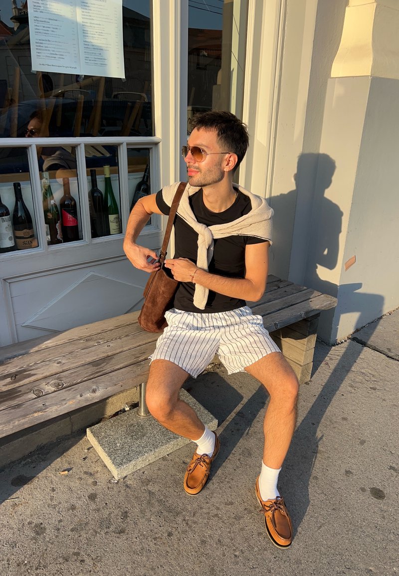 Uma pessoa está sentada num banco de madeira, vestindo uma t-shirt preta, calções às riscas, sapatos castanhos e um suéter leve pousado sobre os ombros.