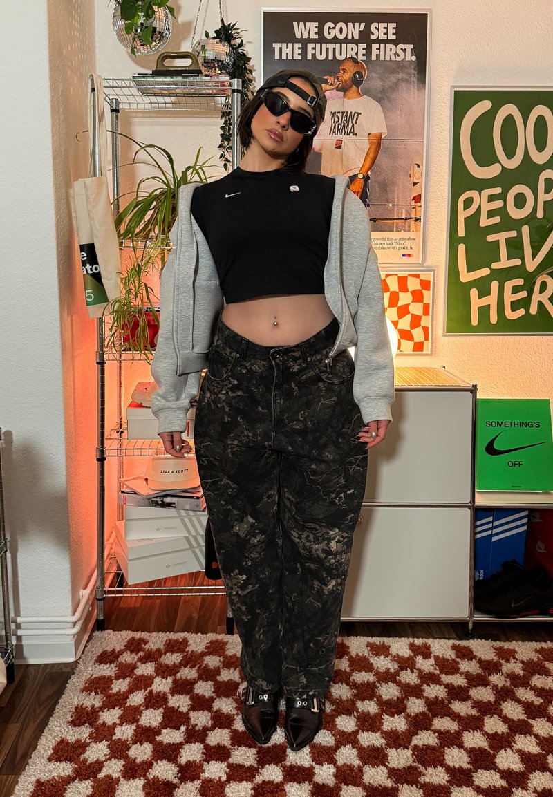 Top court noir, sweat à capuche oversize gris, pantalon foncé à motifs et bottines noires. Des lunettes de soleil et une casquette complètent le look urbain.