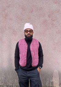 Ροζ βελούδινο γιλέκο πάνω από ένα μαύρο πουλόβερ, φορεμένο με ένα ανοιχτό ροζ σκουφάκι και σκούρα γυαλιά ηλίου. Μαύρο παντελόνι με τσέπες, χαλαρός αστικός στυλ.