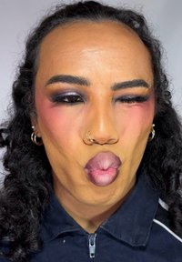 Person med lockigt hår, djärv ögonmakeup, näsringar och örhängen som gör en lekfull min med puckered läppar och ett öga stängt.