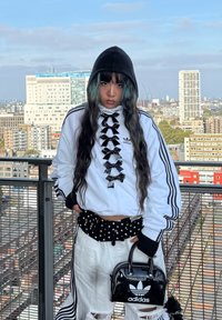 White jacket adorned with black bows, featuring three black stripes. It is paired with a black polka dot belt, distressed white trousers, and a shiny black handbag. The setting is urban.