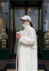 Biely kožušinový kabát s vysokým golierom a opaskom v páse, v kombinácii so svetlou čiapkou. Drží červený papierový pohár, stojí pri výklade obchodu.