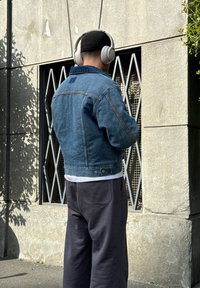 Chaqueta de denim con un lavado azul, costuras en contraste y un parche cuadrado en la parte trasera; combinada con pantalones sueltos de corte gris oscuro y una camiseta blanca.