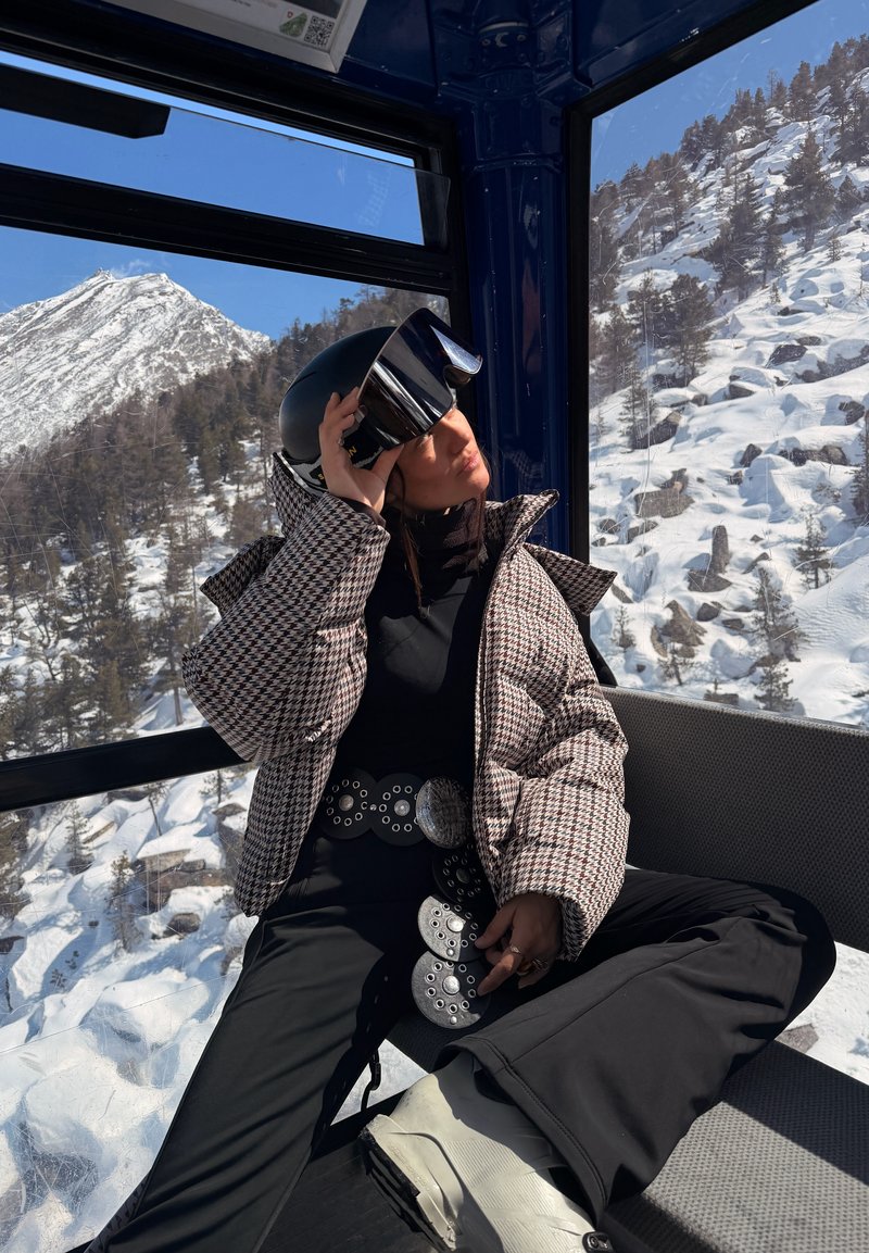 Femme en tenue de ski ajustant son casque à l'intérieur d'une gondole avec une montagne enneigée et des arbres visibles à travers les fenêtres.
