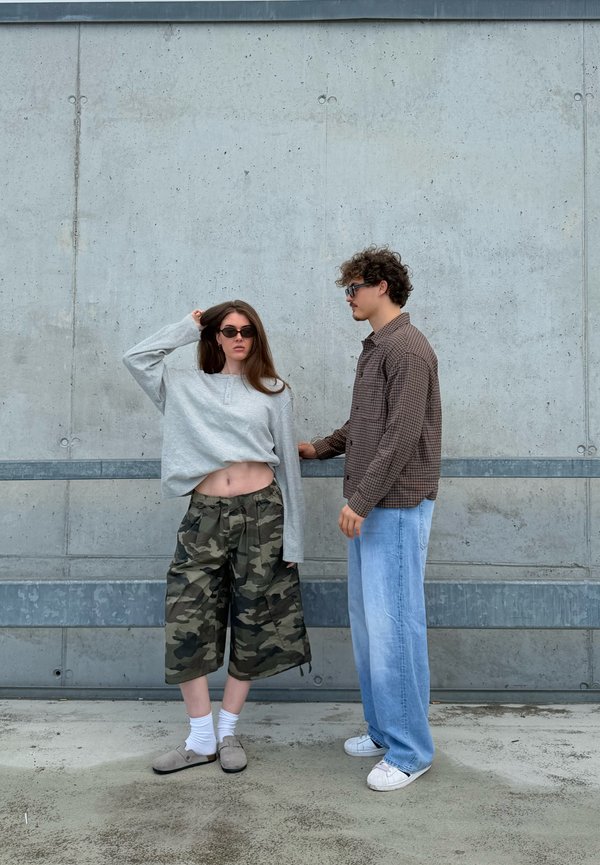 Una donna con occhiali da sole, top corto grigio e pantaloncini mimetici è accanto a un uomo con occhiali da sole, camicia a quadri e jeans blu davanti a un muro di cemento.