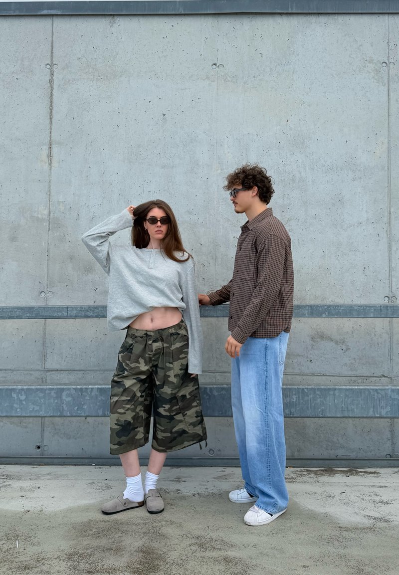 Een vrouw met een zonnebril, een grijze crop top en camouflage shorts staat naast een man met een zonnebril, een geruit overhemd en blauwe jeans tegen een betonnen muur.