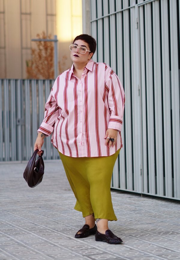 Person with short hair wearing oversized pink striped shirt and green pants, holding a brown bag, standing on tiled sidewalk near metal fence.