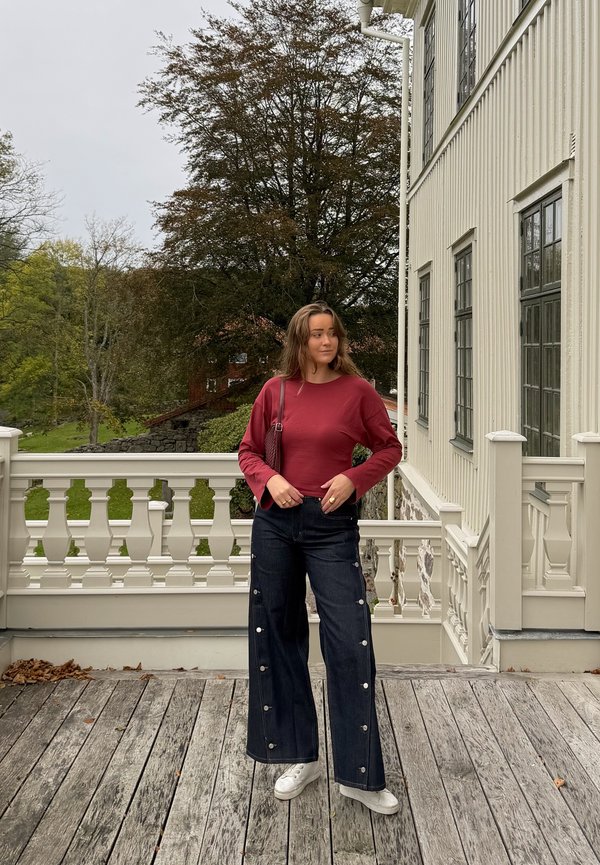 Una mujer con un top rojo y unos vaqueros de pernera ancha posa en una terraza de madera junto a una casa blanca, con árboles detrás.