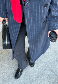 Manteau rayé foncé avec une écharpe rouge, jeans évasés noirs et mocassins noirs. Porte un sac à main noir et un gobelet de voyage. Surfaces texturées visibles.