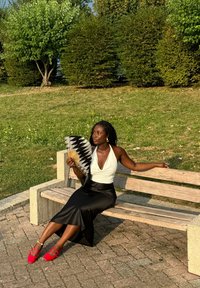 Une femme est assise sur un banc en bois, portant un haut halter blanc et une jupe en satin noire, tenant un éventail à motifs noir et blanc. Des chaussures rouges complètent le look.