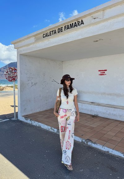 Edificio blanco con el letrero "Caleta de Famara". Persona con camiseta blanca y pantalones de pierna ancha con estampado se encuentra en un área de loseta, usando gafas de sol y un sombrero.
