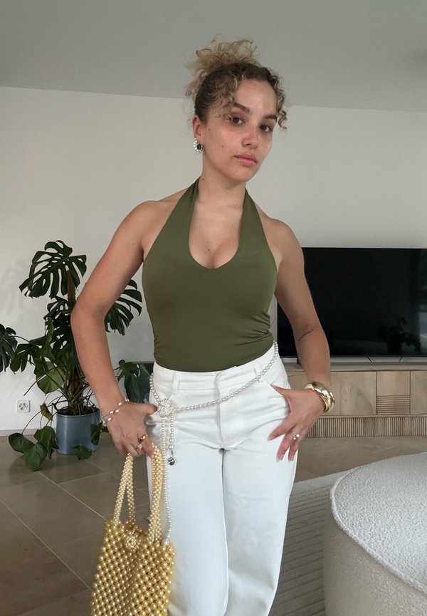 Femme aux cheveux bouclés portant un haut dos nu vert et un pantalon blanc, tenant un sac à main en perles, debout dans un salon moderne.