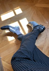 Zwarte leren schoenen met een glanzende afwerking, voorzien van veters en een subtiel ontwerp, gecombineerd met grijs gestreepte broek op een houten vloer.