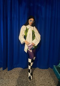 Top verde com lantejoulas sobre uma camisa branca com uma gravata, leggings roxas, botas com padrão xadrez preto e branco, segurando uma pequena mala roxa.