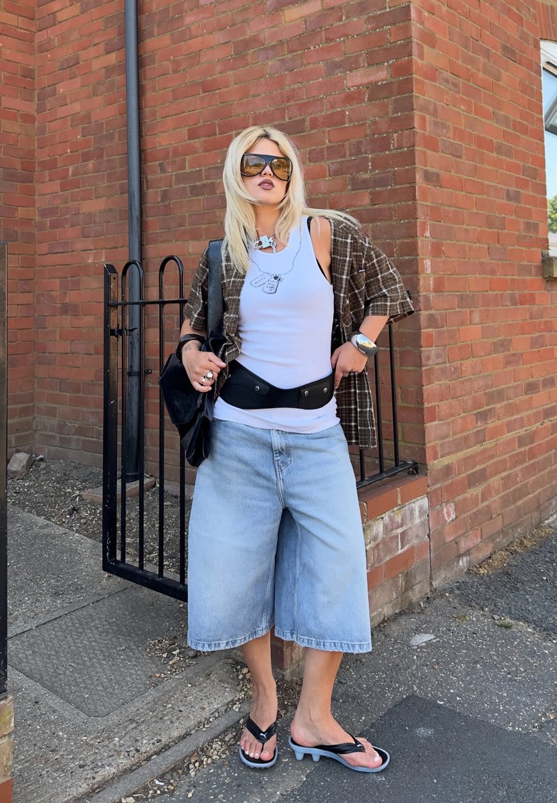 Femme blonde portant un short en jean surdimensionné, un débardeur blanc, une chemise à carreaux, des lunettes de soleil et des sandales noires à talons, debout près d'un mur en briques et d'une porte noire.