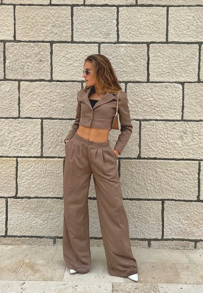 Blazer corto marrón con un solo botón, combinado con pantalones marrones holgados. Tela suave, ligero brillo y bolsillos grandes. Zapatos blancos.