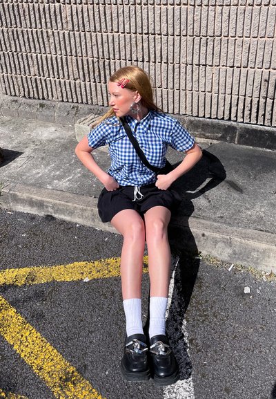 Modelo vistiendo una camisa de cuadros azul y blanca, pantalones cortos negros, calcetines blancos y zapatos negros gruesos con detalles plateados, sentado sobre el pavimento.