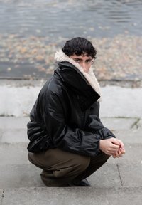 Jong persoon die op betonnen trappen bij het water hurkt, draagt een zwart jack met een grote beige schapenvachtkraag en kijkt recht vooruit.