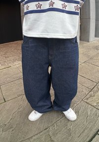 Διακριτές τζιν παντελόνες σε σκούρο μπλε με ορατές ραφές, συνδυασμένες με λευκά αθλητικά παπούτσια. Το ύφασμα έχει απαλή υφή και είναι ελαφρώς φθαρμένο.