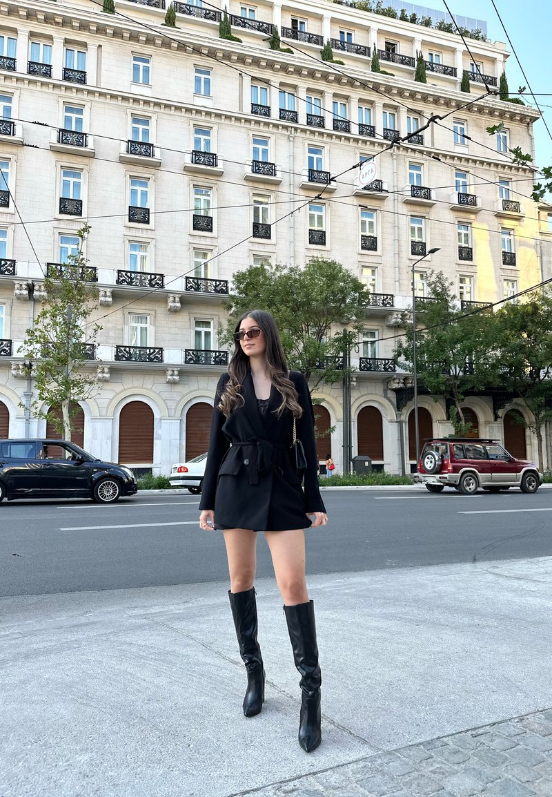 Jeune femme en robe blazer noire et bottes hautes se tient sur le trottoir de la ville avec un bâtiment de style européen classique et des voitures garées derrière.