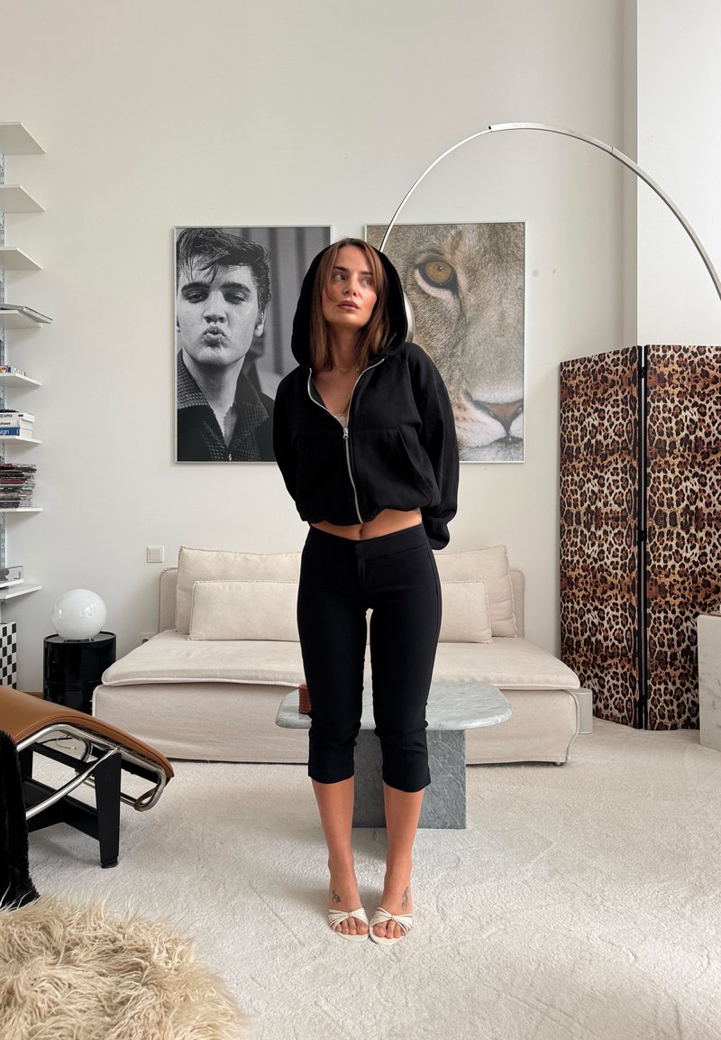 Femme en hoodie courte noire et pantalon se tient pieds nus dans un salon moderne avec un canapé beige, une table en marbre, un paravent à imprimé léopard et une grande œuvre murale.