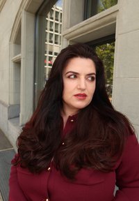 Kobieta z długimi ciemnymi włosami i czerwoną szminką, ubrana w burgundową bluzkę, stoi na zewnątrz w pobliżu kamiennego budynku z oknami odbijającymi miejską panoramę.