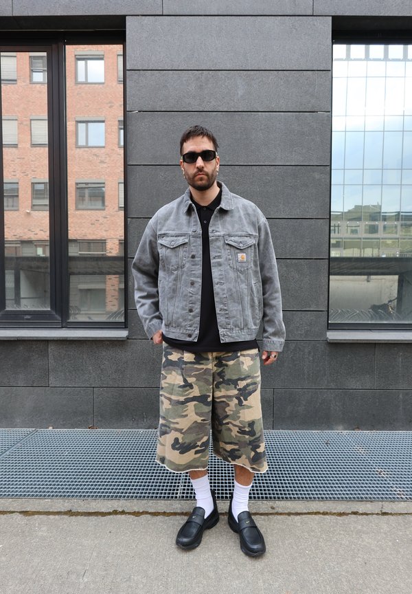 Bărbat purtând ochelari de soare închiși la culoare, jachetă gri din denim, tricou negru, pantaloni scurți camuflați, șosete albe și încălțăminte neagră stă pe trotuarul urban.