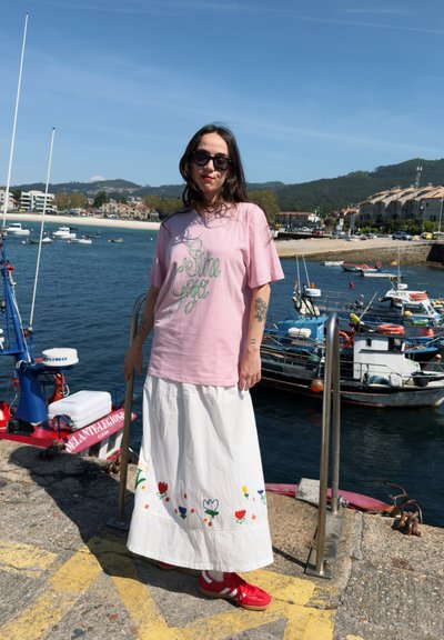 Camiseta rosa con texto en cursiva verde, combinada con una falda blanca con bordados florales. Zapatillas rojas completan el look. Fondo de botes y agua.