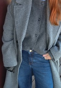 Osoba ubrana w długi szary wełniany płaszcz, szary sweter zapinany na guziki, niebieskie dżinsy, czarna skórzana rękawiczka na lewej ręce, prawa ręka w kieszeni.