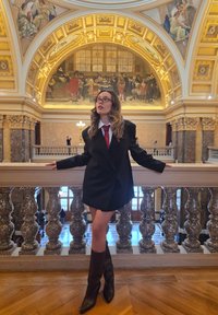 Czarna, oversize'owa marynarka na białej koszuli i czerwonym krawacie; modelka nosi brązowe, skórzane kozaki sięgające do kolan. Ozdobne wnętrze z marmurowymi elementami.