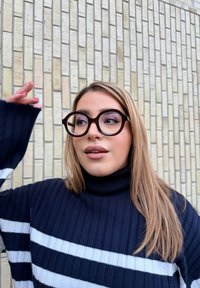 Στρογγυλά, σκούρα καφέ γυαλιά με παχιές γωνίες, φορεμένα από ένα άτομο με ναυτική ριγέ πουλόβερ με λαιμόκοψη, μπροστά από έναν υφασμένο φωτεινό τοίχο από τούβλα.