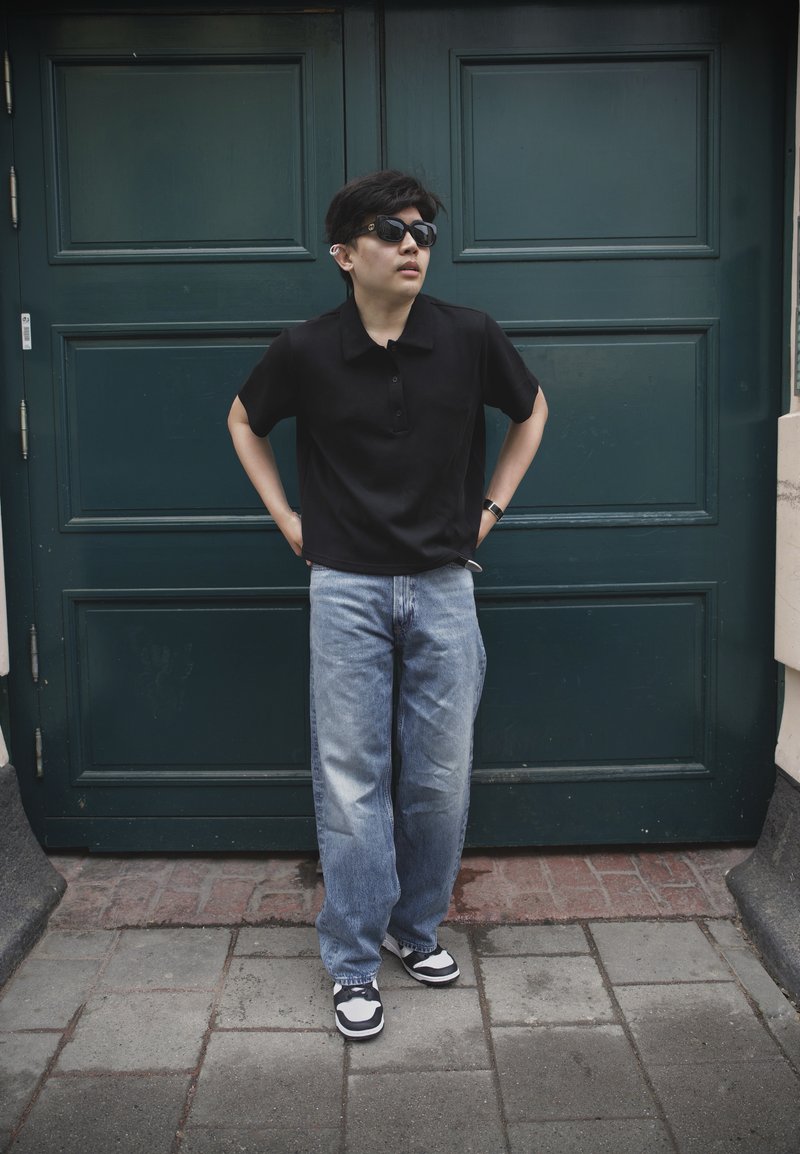 Personne portant des lunettes de soleil, un polo noir, un jean bleu ample et des baskets, debout devant une porte vert foncé, les mains sur les hanches.