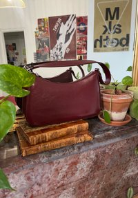 Un sac à main en cuir marron repose sur deux livres anciens sur une surface en marbre, entouré de plantes en pot et d'affiches sur un mur blanc.