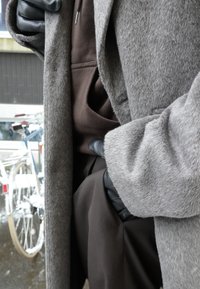 Personne portant un manteau gris, un sweat à capuche marron, et des gants en cuir noir avec les mains dans les poches, debout près de vélos à l'extérieur.
