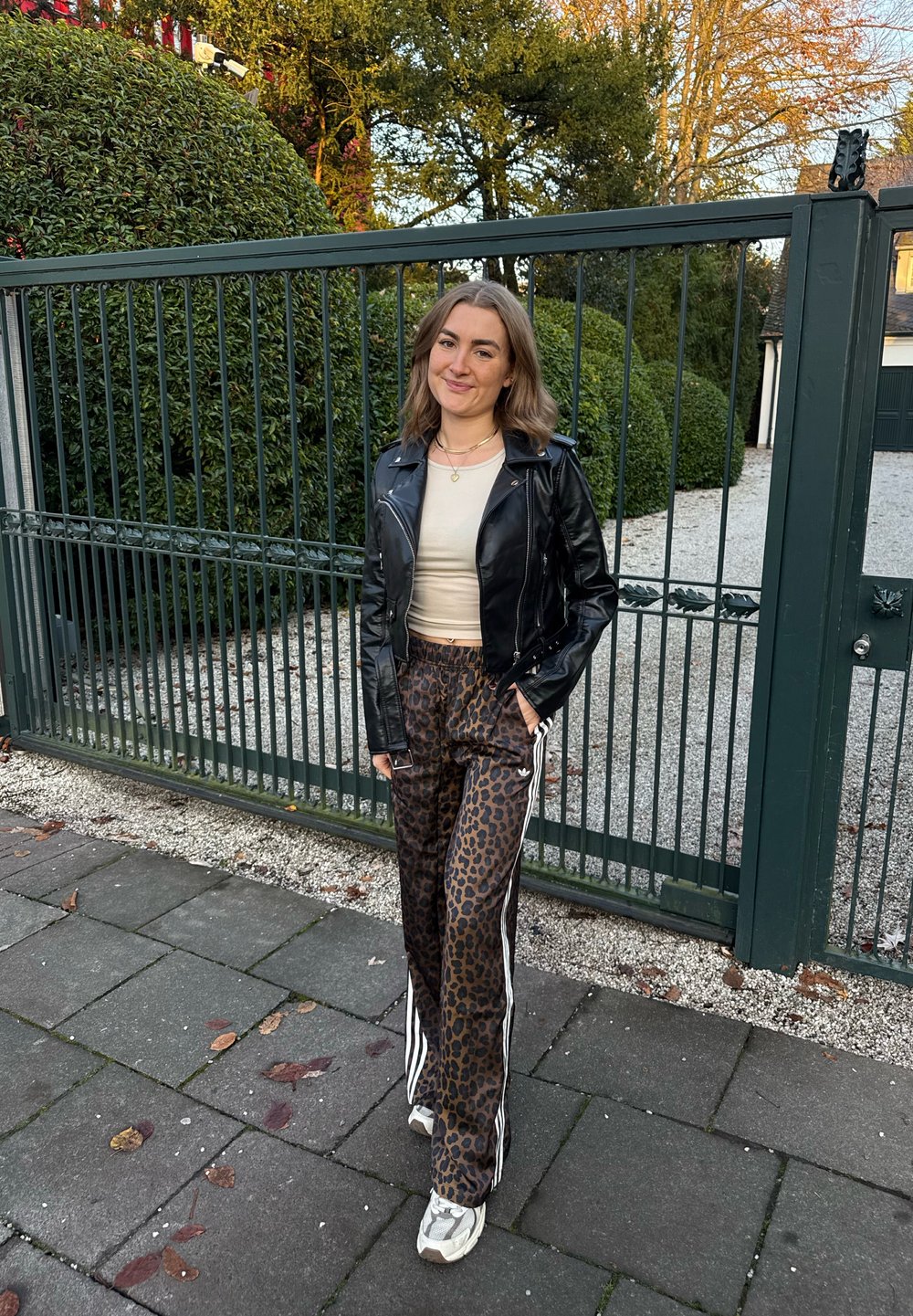 Chaqueta de cuero negra, camiseta ajustada beige, pantalones marrones con estampado de leopardo y rayas blancas a los lados, y zapatillas grises. De pie cerca de un área cerrada con rejas.