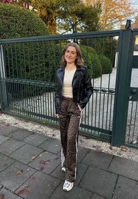Chaqueta de cuero negra, camiseta ajustada beige, pantalones marrones con estampado de leopardo y rayas blancas a los lados, y zapatillas grises. De pie cerca de un área cerrada con rejas.