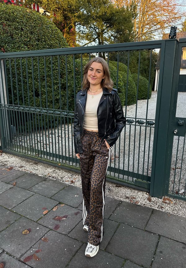 Veste en cuir noire, haut ajusté beige, pantalon marron à imprimé léopard avec des bandes latérales blanches, et baskets grises. Debout près d'une zone clôturée.